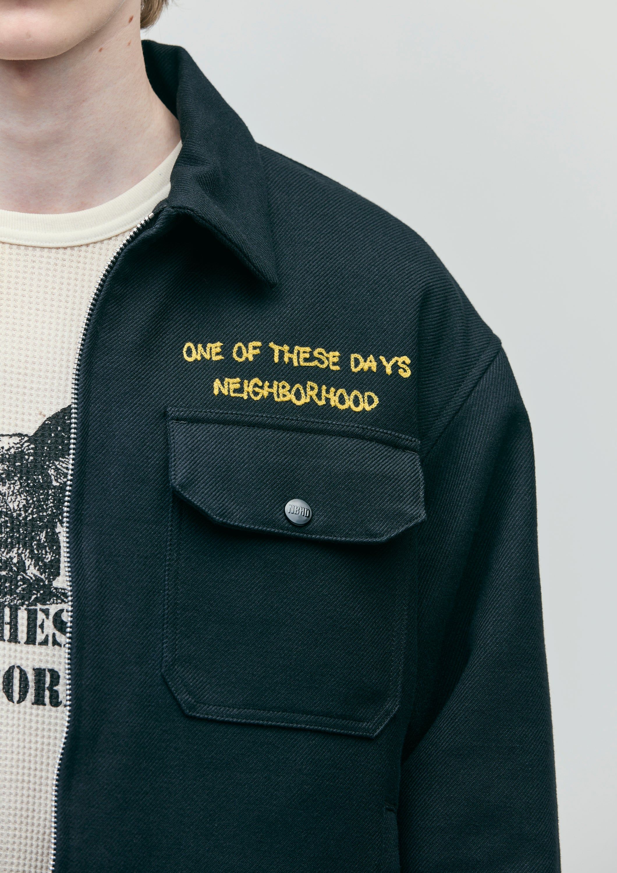 A contemporary streetwear-inspired overshirt featuring utilitarian design elements with embroidered text branding. The piece combines minimalist aesthetics with functional details, layered over a graphic tee, creating a casual yet intentional visual statement.