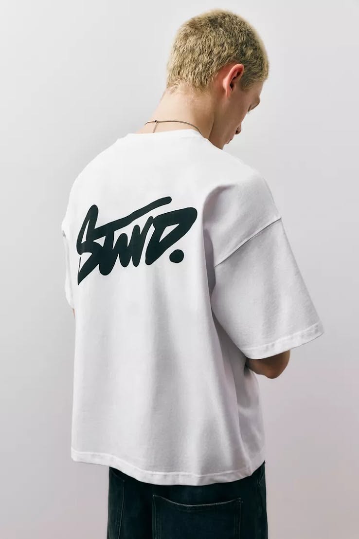 This image features a close-up of a white t-shirt with a bold, black graphic print on the back. The design utilizes a stylized, brushstroke-like script that conveys a sense of edgy, contemporary streetwear branding.