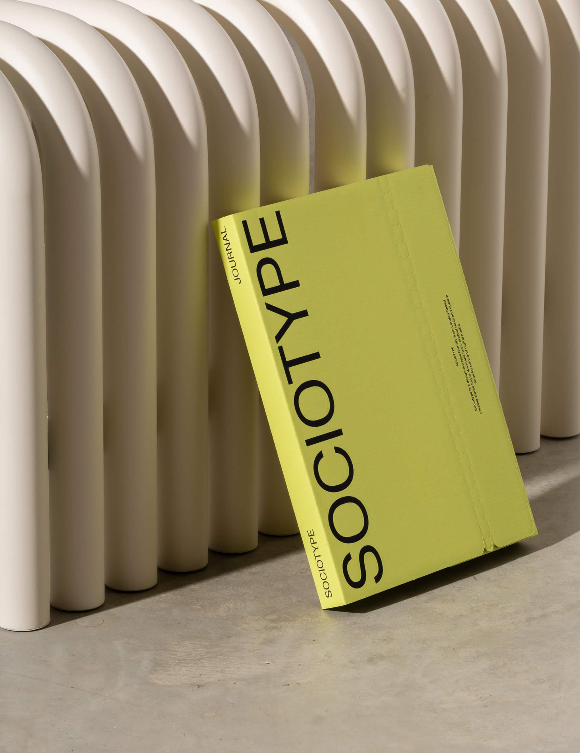 The image presents a clean, minimalist study in form and texture, contrasting the smooth, vertical architectural elements with the solid, tactile presence of a brightly colored book. The visual language emphasizes subtle shadows and clean lines, creating a sophisticated and serene atmosphere.