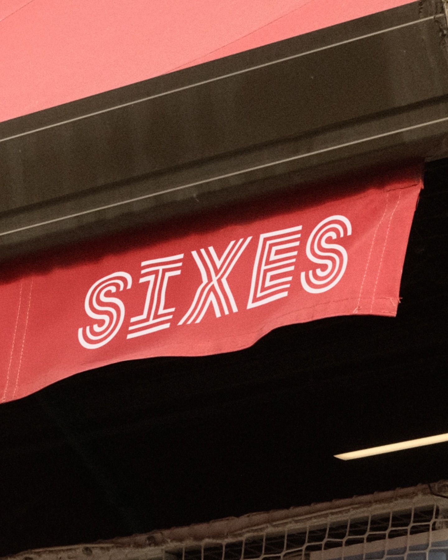 The image features bold, large typography set against a vibrant pink fabric banner hanging beneath a dark structure. The design relies on high contrast between the bright lettering and the deep shadows, creating a striking and attention-grabbing visual statement.