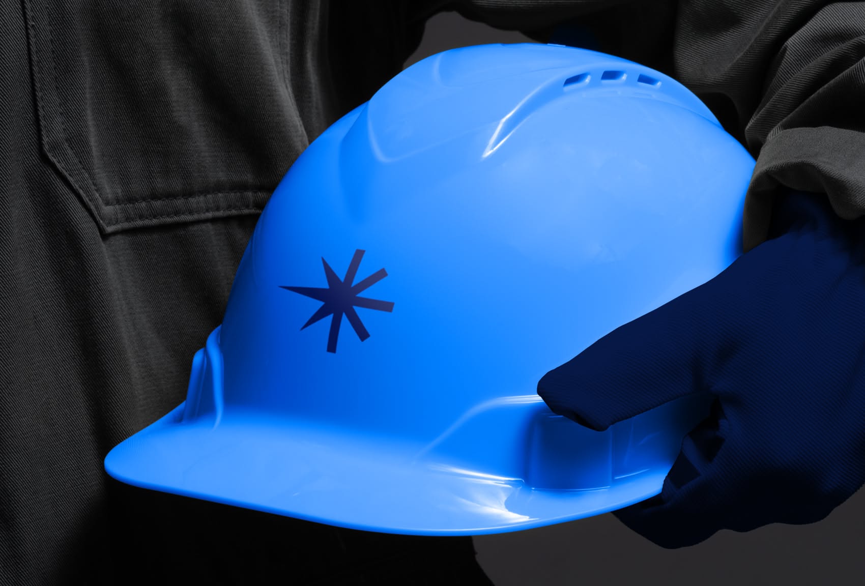 This image is a clean product shot emphasizing the texture and color of a bright blue safety hard hat being held by a dark glove. The visual language relies heavily on stark contrast between the smooth plastic, the matte fabric of the hand, and the deep shadows, conveying a sense of professional utility and protection.