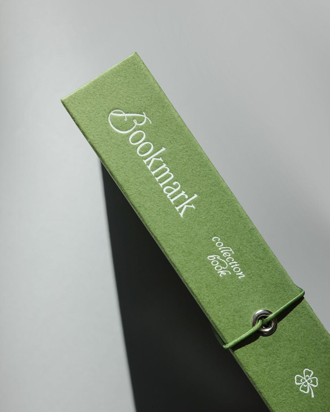 This image features a close-up of a rectangular, olive-green fabric item, likely a bookmark, with elegant white text printed on it. The design emphasizes simplicity, natural texture, and understated sophistication through its muted color and clean typography.