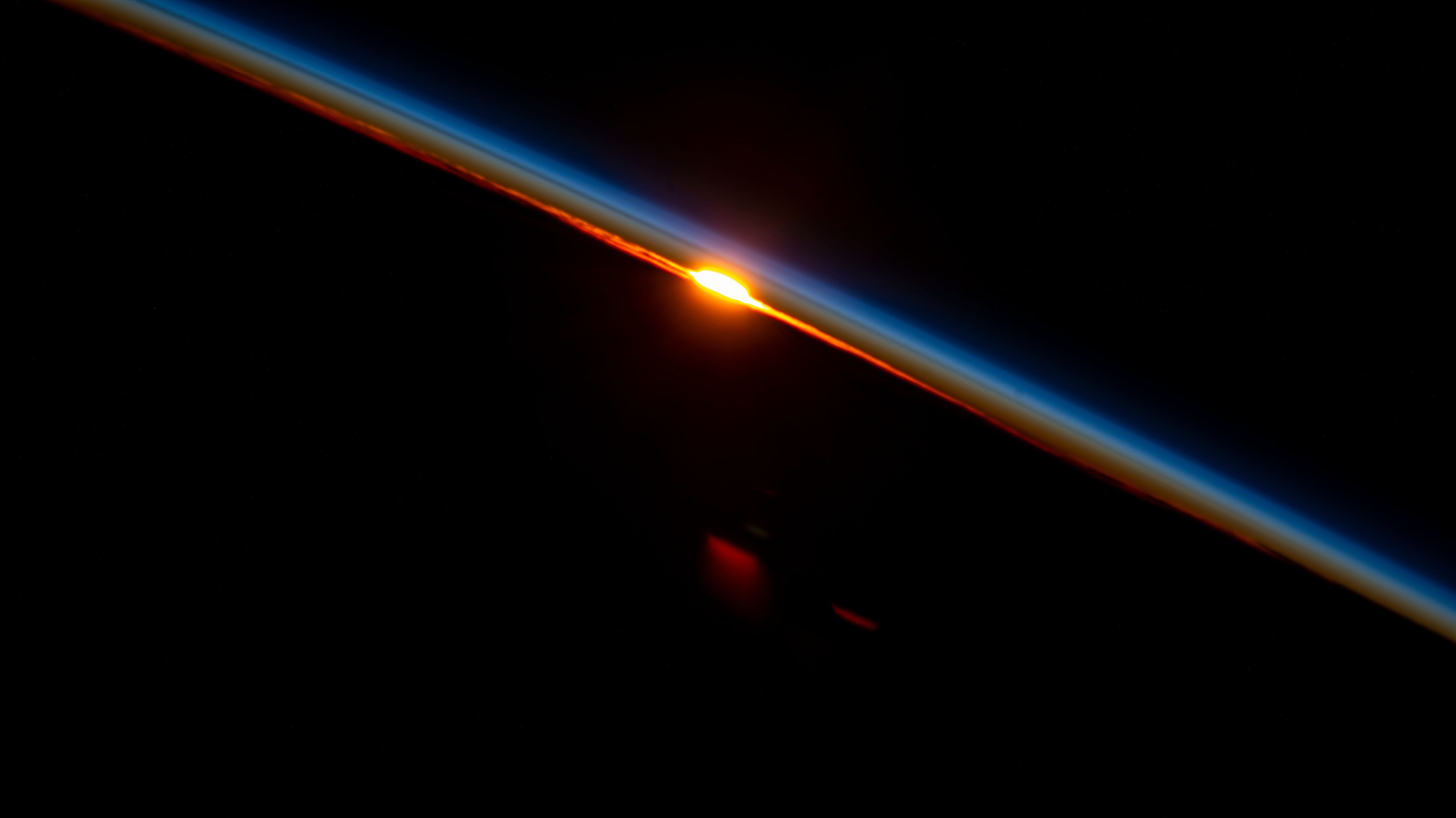 This image captures a dramatic view of the Earth's limb against the blackness of space, highlighting the thin blue atmosphere and a brilliant solar flare or atmospheric glow. The visual language is stark, emphasizing the contrast between light and deep shadow.