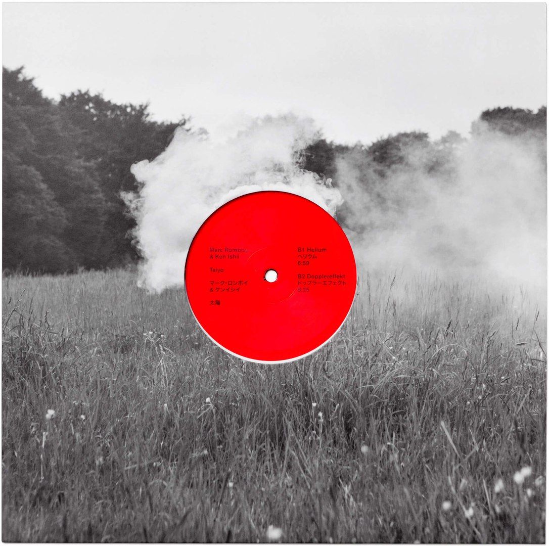 This is a high-contrast, monochrome photograph featuring a vinyl record centered within a cloud of smoke or fog. The composition is stark and dramatic, emphasizing texture and the ephemeral nature of the smoke against the solid, circular form of the disc.