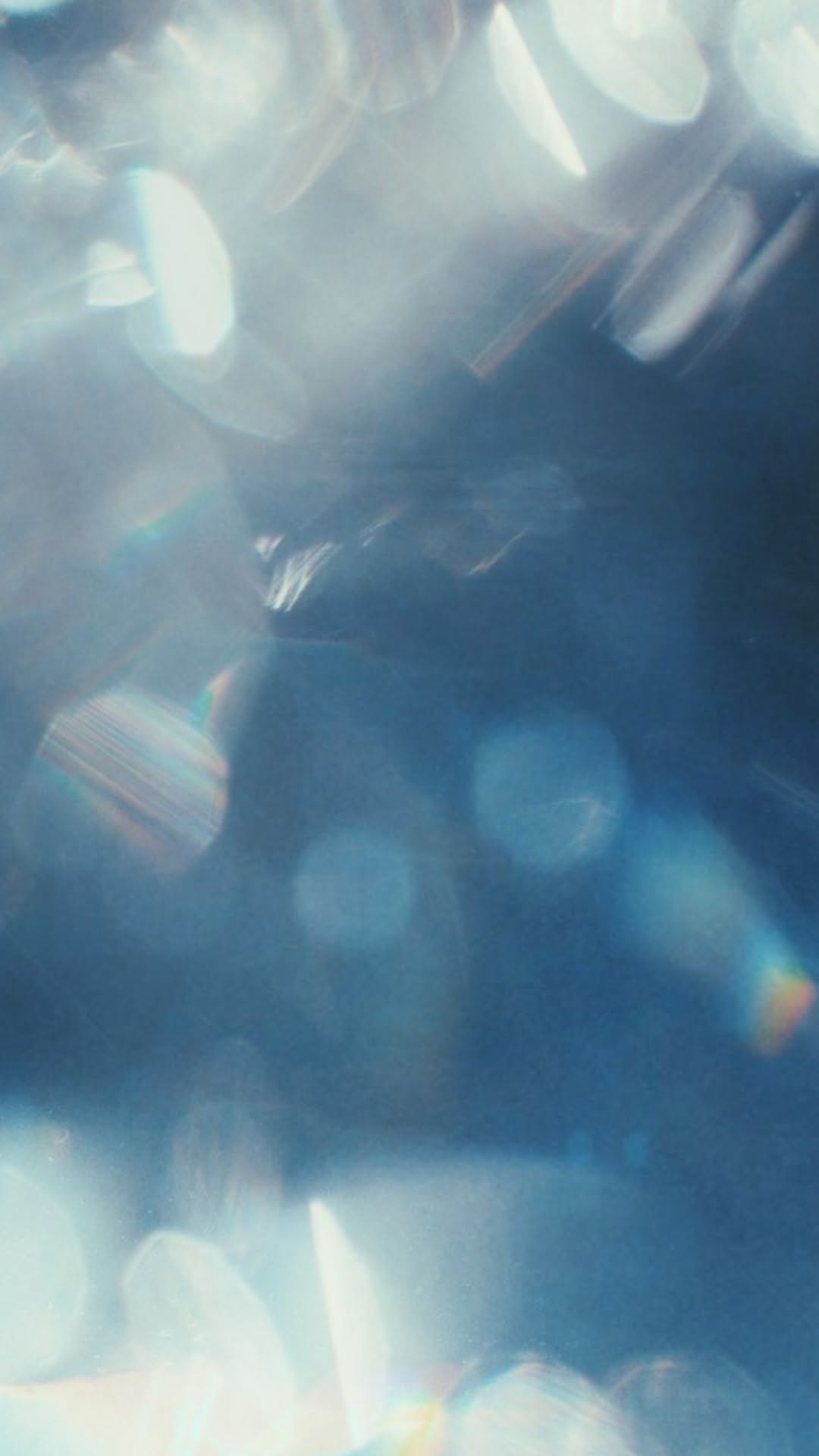 This image is an abstract, out-of-focus close-up featuring bright light flares and bokeh effects against a dark blue background. The visual language is characterized by soft focus, high contrast between light and shadow, and a sense of movement or sparkle.