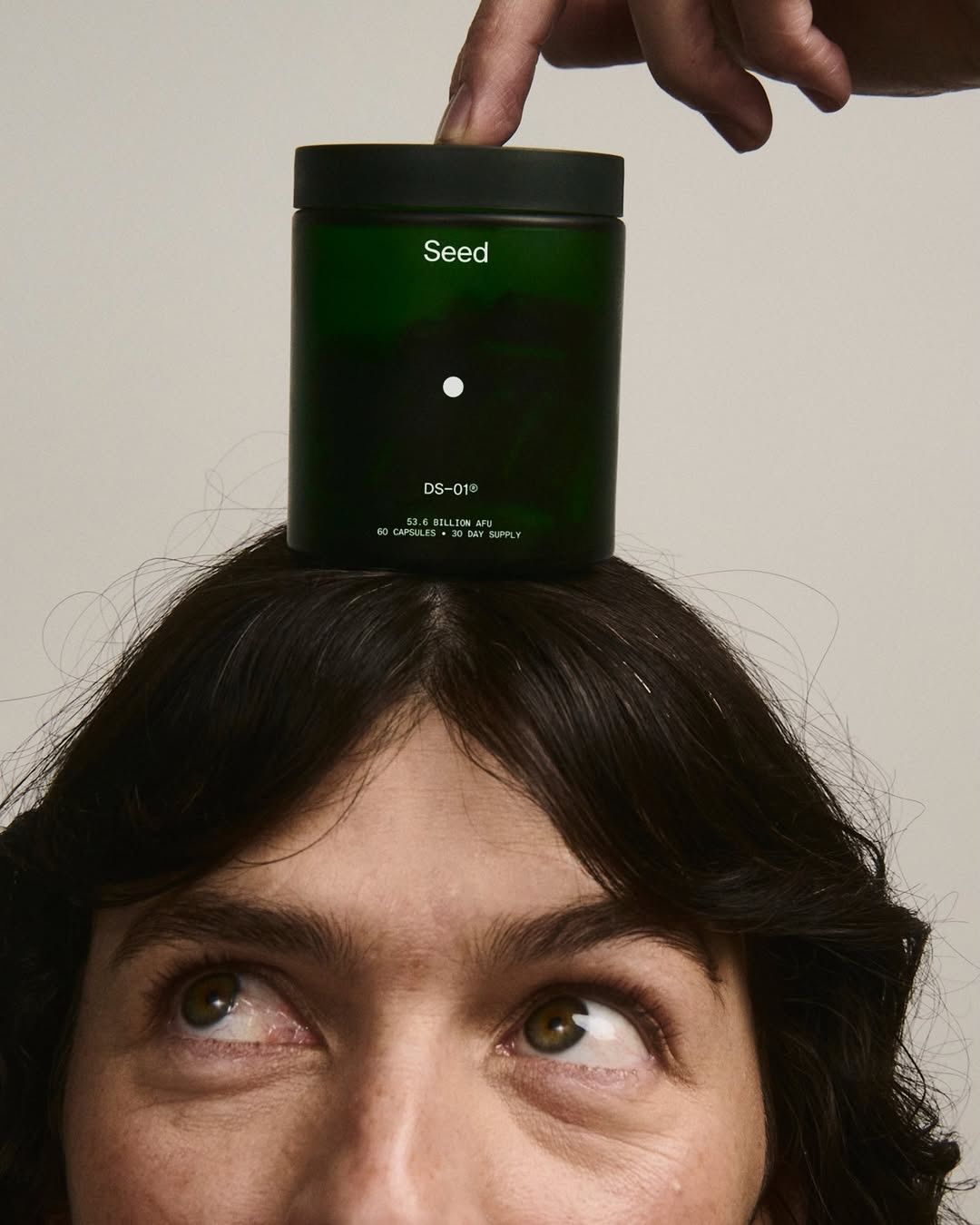 This is a high-contrast, minimalist product photograph that positions a dark green wellness jar above a person's face. The visual language emphasizes naturalness and purity through simple forms and soft, even lighting, creating a serene yet engaging composition.