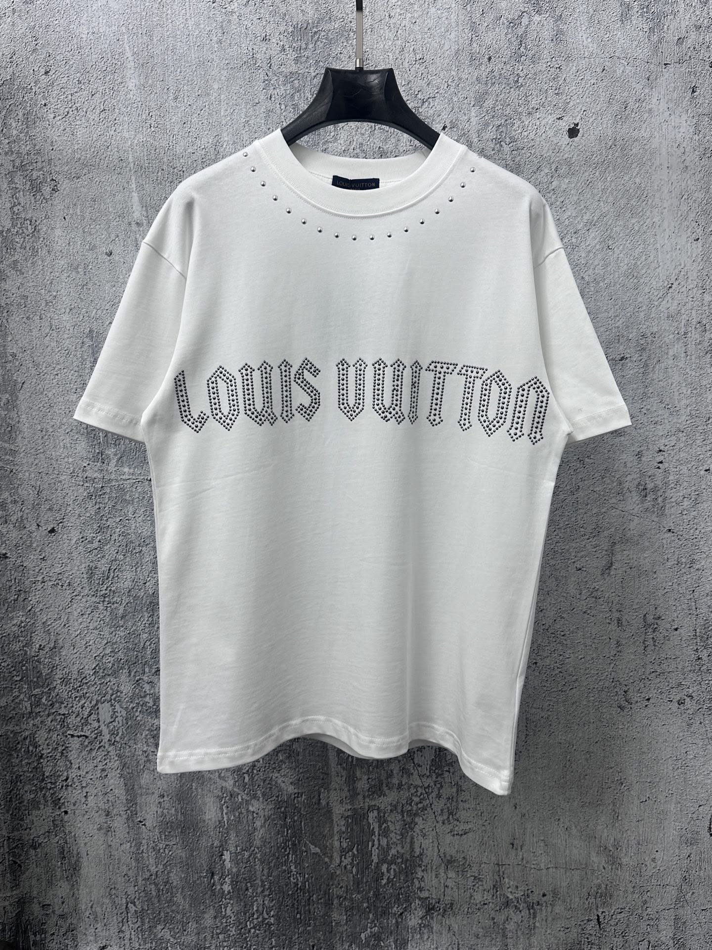 This image showcases a minimalist white t-shirt featuring a subtle, dotted typography print for the Louis Vuitton logo. The design utilizes simple clean lines and high-contrast texture against a rugged, monochromatic gray background to emphasize quiet luxury and brand detail.
