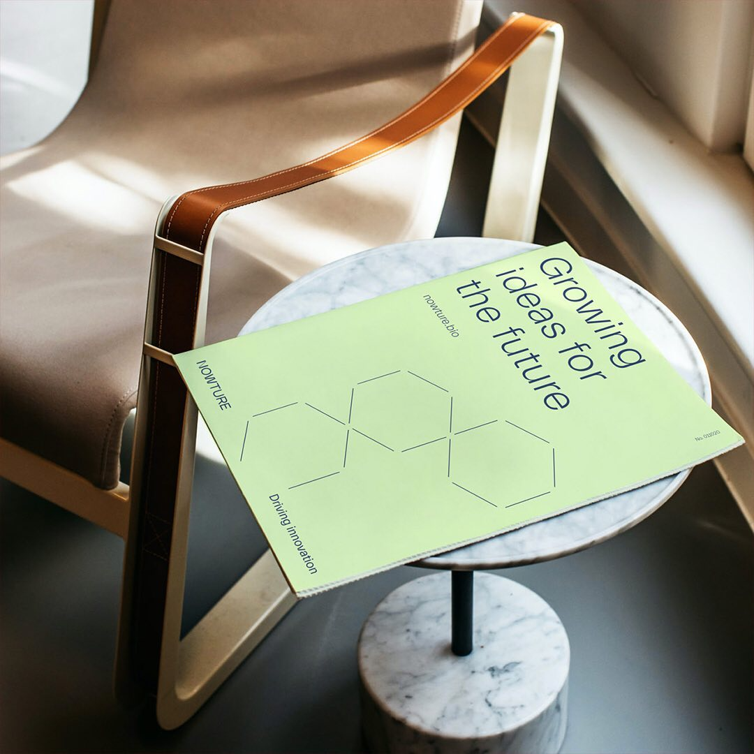 A minimalist, contemporary styled shot featuring a lime green publication with geometric hexagon illustrations placed on a marble side table next to a mid-century modern chair with cognac leather straps. The composition emphasizes clean lines, natural light, and sophisticated material contrasts in a curated interior setting.