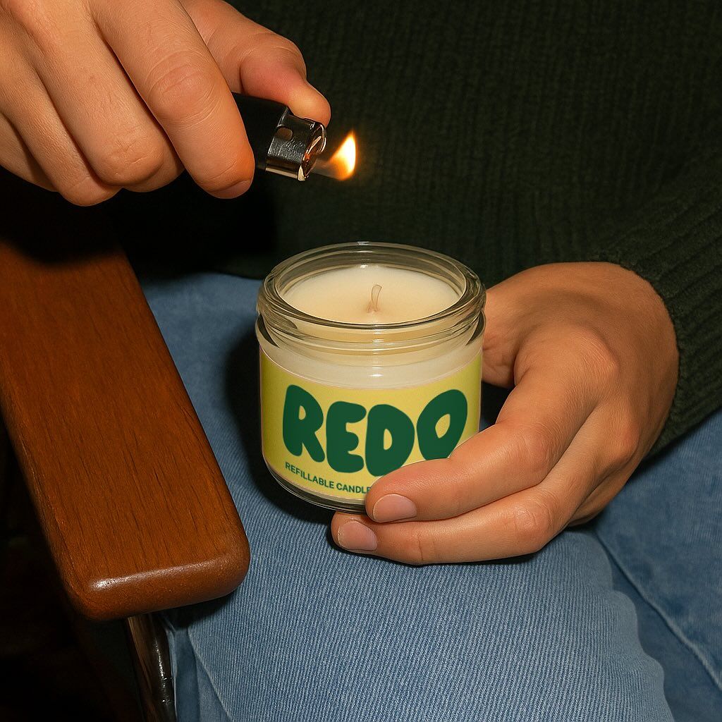 A lifestyle product photography shot featuring a hand-held candle with bold branding being lit by a lighter. The composition captures an intimate moment of product interaction against a soft, neutral background with warm and cool tonal contrasts. The image conveys authenticity through natural hand positioning and candid lighting.