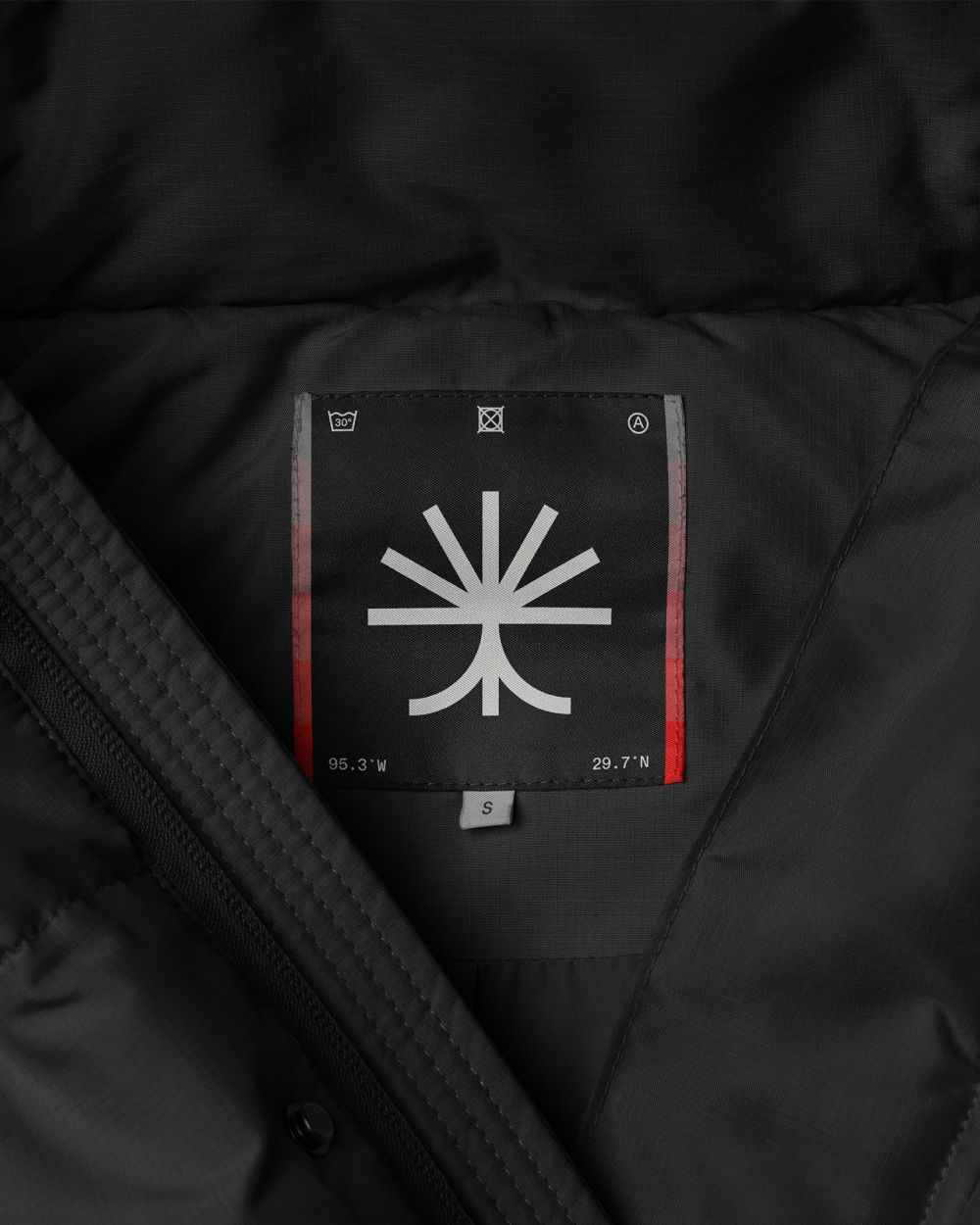 This image presents a close-up view of a dark, technical garment, focusing on a branded patch or label detail. The visual language is minimalist and functional, relying on stark contrast between the matte black fabric and the white/light gray graphic. The overall feel is precise, technical, and premium.