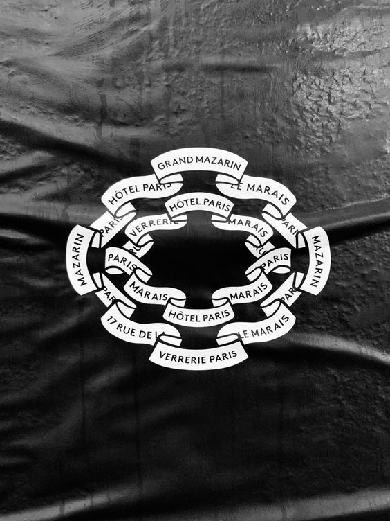 A sophisticated black and white emblem featuring a circular ribbon banner design with Parisian hotel and landmark names arranged in a decorative wreath pattern. The design combines classical heraldic elements with Art Deco sensibilities, creating an elegant, vintage aesthetic that evokes early 20th-century luxury branding.