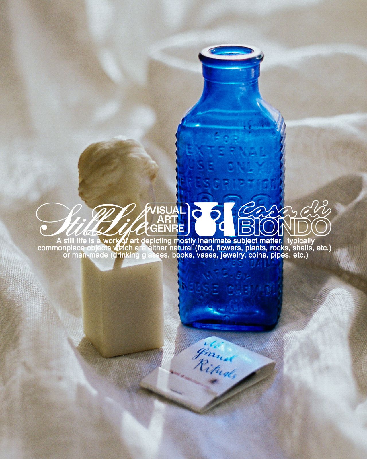 This is a still life photograph featuring a vibrant blue glass bottle alongside a pale, sculpted object, set against a soft, draped white or cream fabric. The composition emphasizes texture contrast and the interplay between smooth glass and matte material, creating a quiet, contemplative mood.